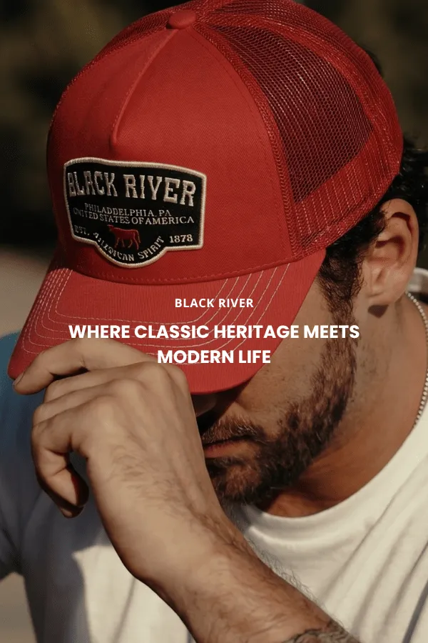 En mann sitter på en trekasse i et studio og bærer en klassisk brun cowboyhatt i filt med bred brem, en mørk denimskjorte og jeans.