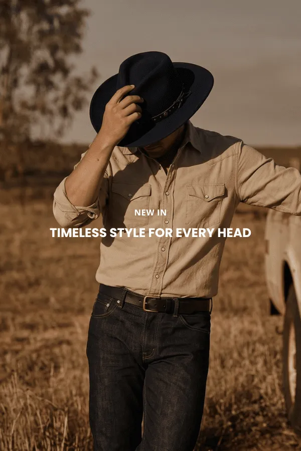 En mann sitter på en trekasse i et studio og bærer en klassisk brun cowboyhatt i filt med bred brem, en mørk denimskjorte og jeans.