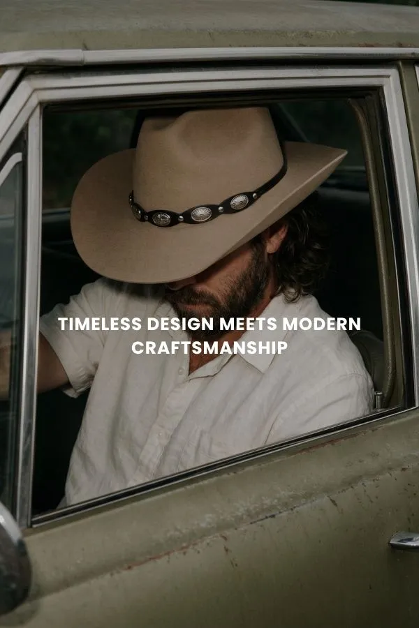 En mann sitter på en trekasse i et studio og bærer en klassisk brun cowboyhatt i filt med bred brem, en mørk denimskjorte og jeans.